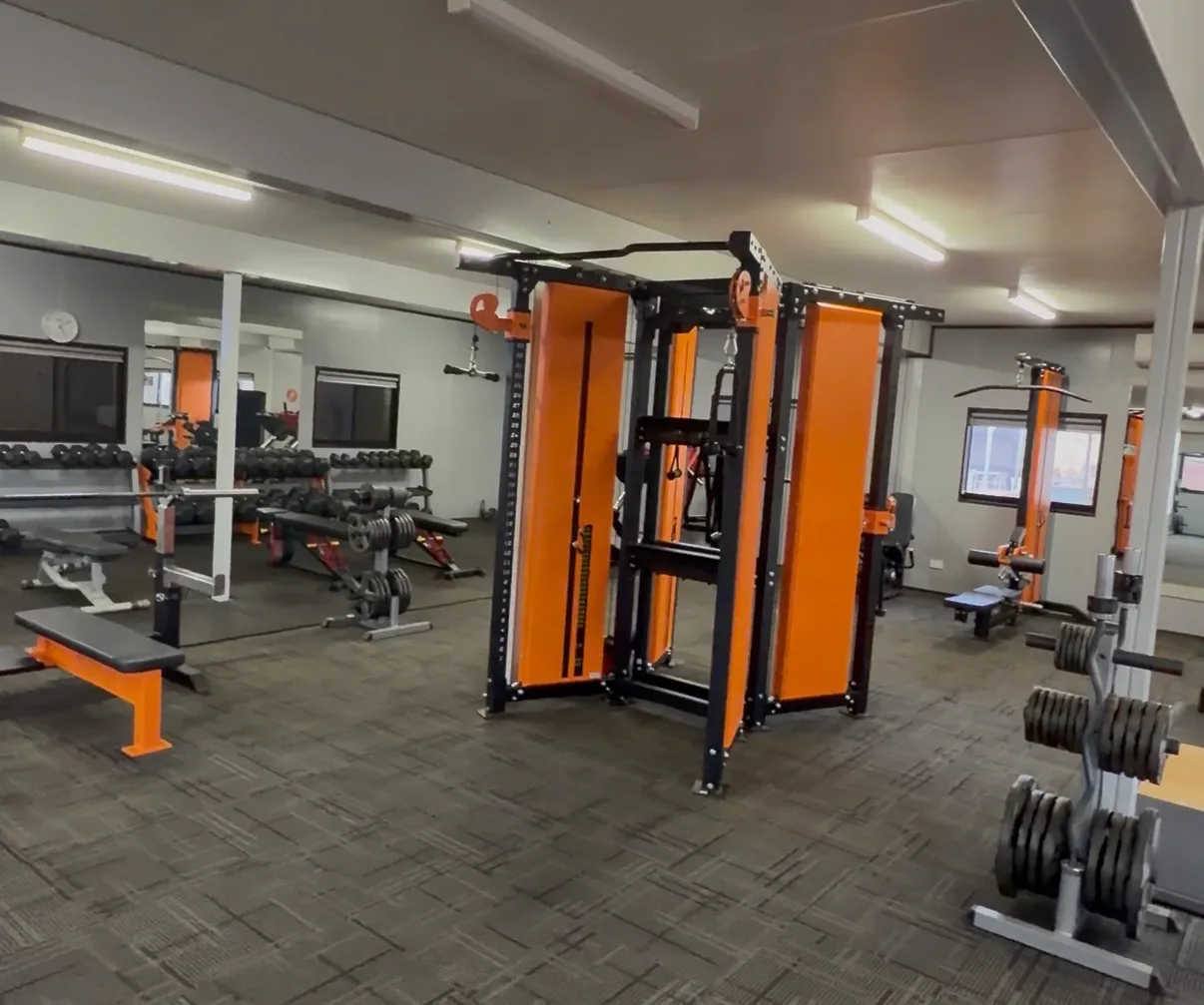 Fitnessraum in einem Bergbaucamp mit orangefarbener Kraftstation, Hantelbank und Kurzhantelablage